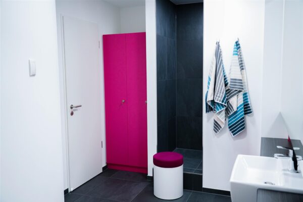 Hygienischer und komfortabler Umkleidebereich in der ESlipo Privatklinik Modern gestalteter Umkleide- und Duschbereich mit pinkem Spind, gefliestem Duschraum, Hockern und gestreiften Handtüchern.